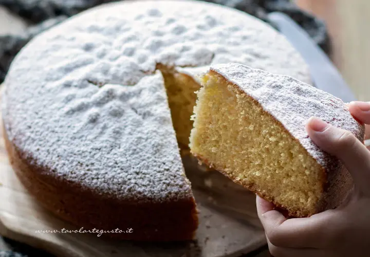 Una fetta di torta margherita ricetta originale, soffice e cosparsa di zucchero a velo, viene sollevata da una mano.