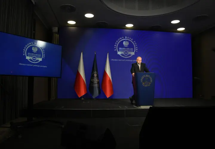 Prezes NBP Adam Glapiński przemawia na tle flag i ekranu z logo banku. Wiron 1m.