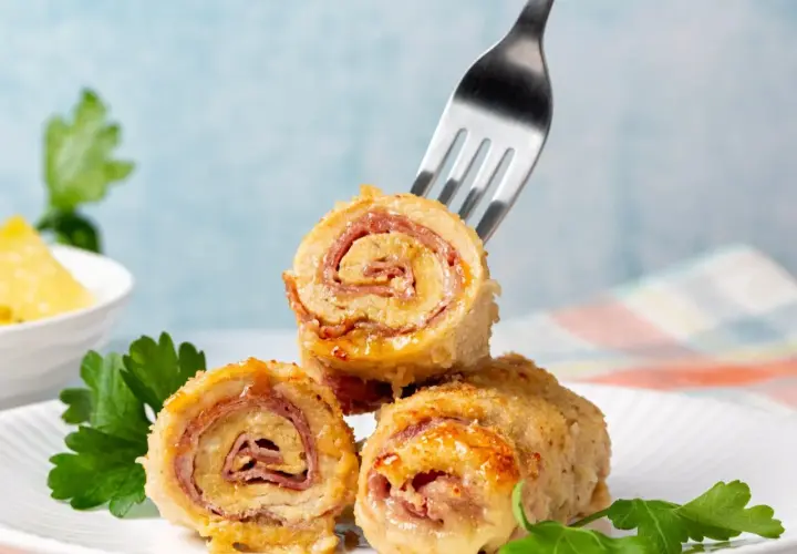 Roladki drobiowe z serem, zawinięte z szynką, prezentują się apetycznie na białym talerzu. Jedna z nich jest nabita na widelec.