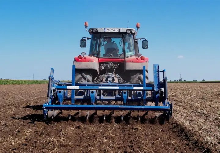 Traktor mit einem blauen Gerät für pfluglose Bodenbearbeitung auf einem Feld unter blauem Himmel.