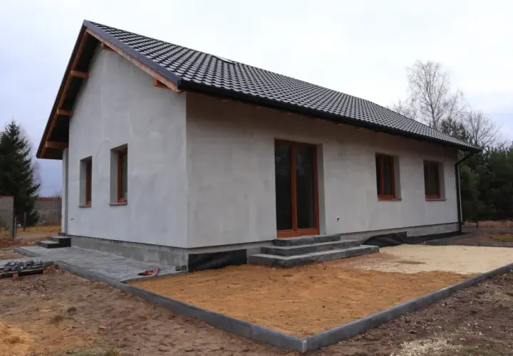 Nowy dom w trakcie budowy, z szarym tynkiem i brązowymi oknami. Wniosek o warunki zabudowy złożony, teren wokół domu przygotowany do dalszych prac.