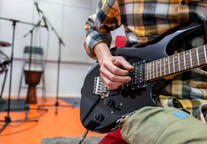 Ręce muzyka grające na czarnej gitarze elektrycznej. W tle stojaki z mikrofonami i bęben. To może być początek nauki, jak zmienić struny w gitarze elektrycznej.