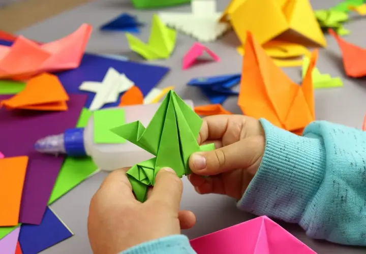 Dziecko tworzy proste origami z kartki a4. Wokół kolorowe figurki i klej.
