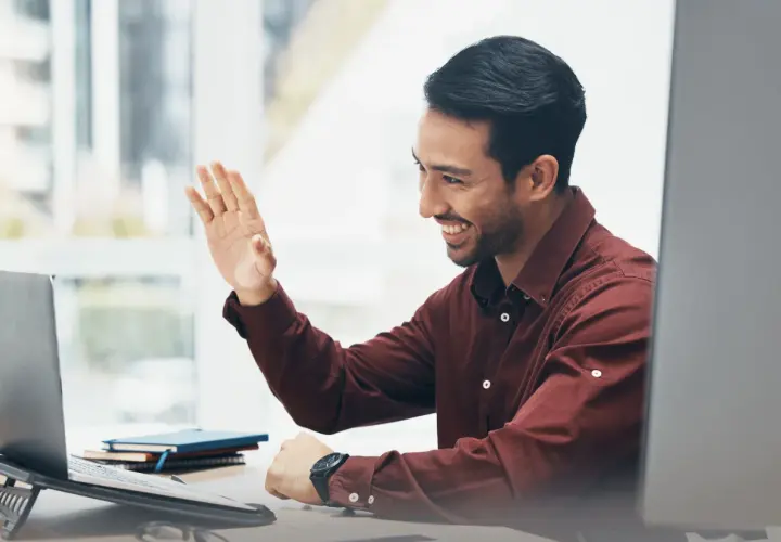 Man waves hello during an online emotional intelligence coaching session, smiling warmly at his laptop.