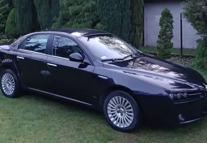 Czarna Alfa Romeo 159 lśni na trawie, otoczona zielenią drzew.