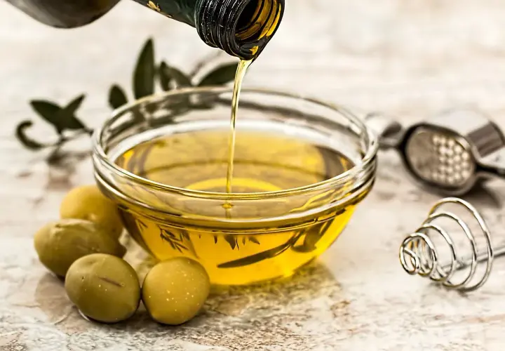 Olio d'oliva versato in una ciotola, pronto per friggere. Olive verdi e un pestello completano la scena.