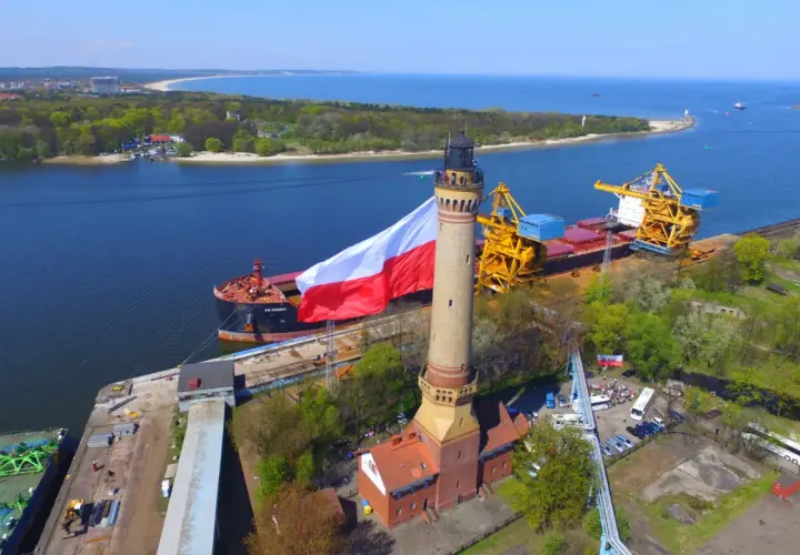 Najwyższa latarnia w Polsce z polską flagą, obok statku i portowych dźwigów.