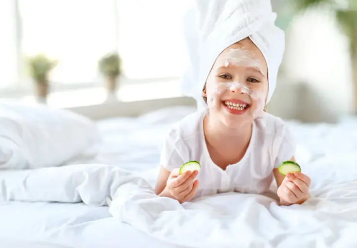 Uśmiechnięta dziewczynka z ręcznikiem na głowie i białą glinką na twarzy cieszy się domowym spa, trzymając w dłoniach plasterki ogórka.