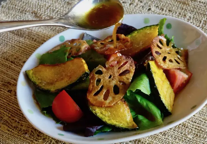 A spoon pours a golden dressing over a salad of roasted pumpkin, lotus root, tomatoes, and greens. The dressing, with a hint of wasabi, adds a zesty kick.