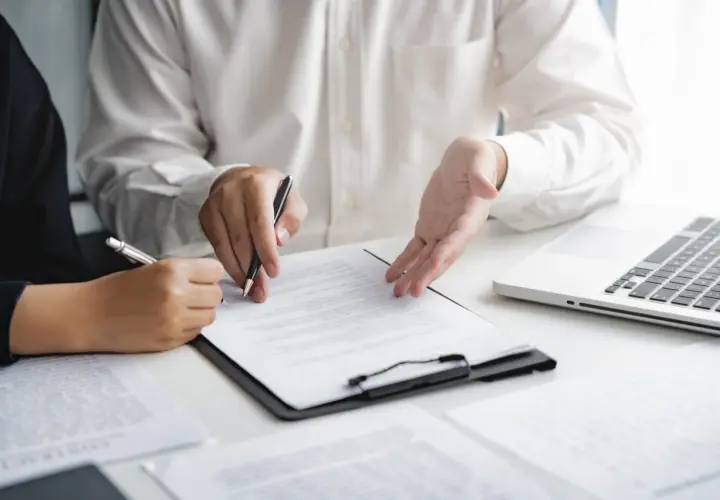 Deux personnes discutent d'un document, l'une signant, l'autre expliquant. Une commission d'intervention est en cours.