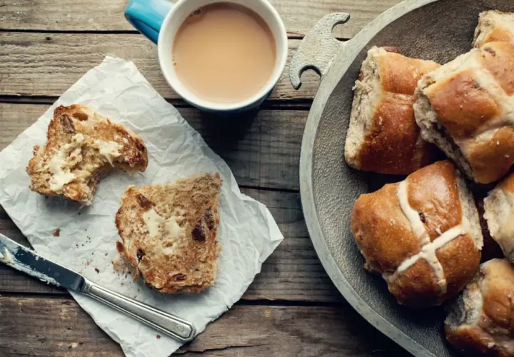 Na drewnianym stole leżą świeże, pachnące hot cross buns, obok kubek kawy i kromka posmarowana masłem.