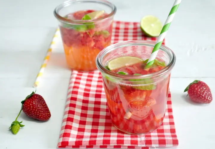 Orzeźwiający drink z truskawkami, miętą i limonką, podany w szklance z zielono-białą słomką. Obok leży świeża truskawka.
