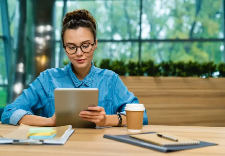 Studentka w okularach korzysta z iPada na studia, siedząc przy biurku z kawą i notatkami.
