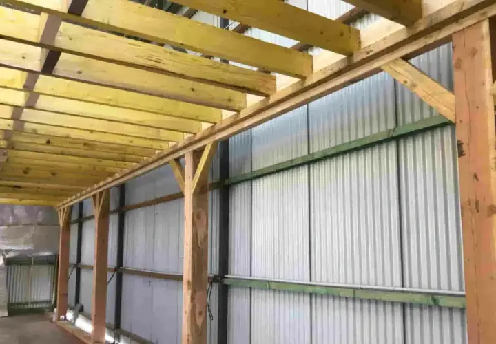 Structure en bois avec madriers ou bastaings soutenant un toit, le long d'un mur en tôle ondulée.