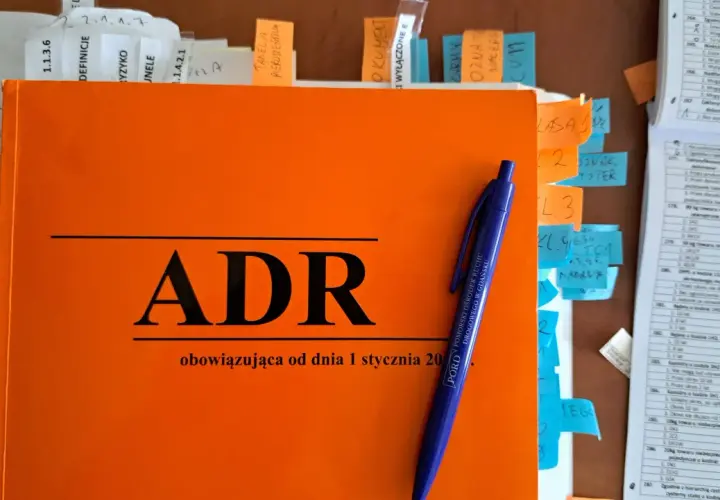 Pomarańczowa książka z napisem "ADR" i niebieski długopis. To materiały do kursu **doradca ADR**.