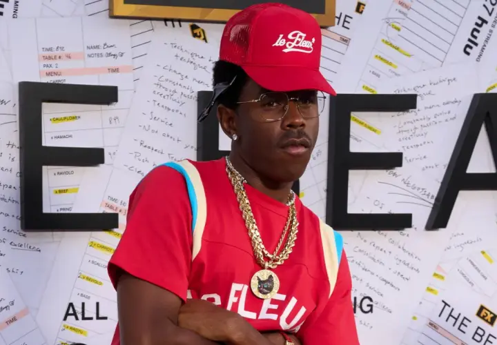 Tyler, The Creator w czerwonej czapce i naszyjnikach, na tle liter "HE" i notatek.