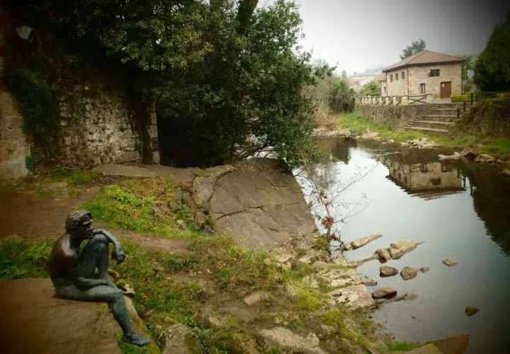 Escultura de hombre pensativo junto a un río, con una casa de piedra reflejada en el agua. Un lugar tranquilo que ver cerca de Liérganes.