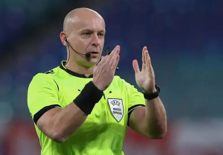 Sędzia piłkarski w żółtej koszulce z emblematem UEFA, z mikrofonem przy uchu, pokazuje gest rękami. Ciekawe, ile zarabia sędzia na takim meczu.