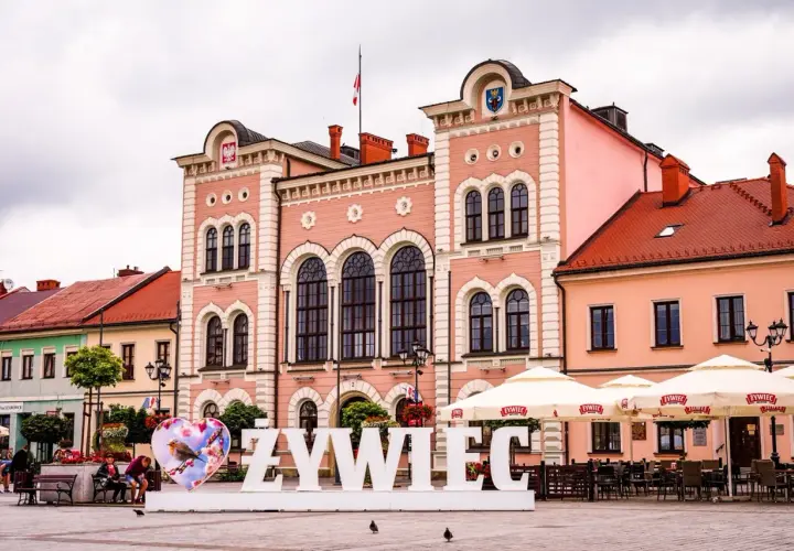 Duże białe litery "ŻYWIEC" na rynku, obok serca z ptaszkiem. W tle różowy ratusz i kawiarniane parasole.