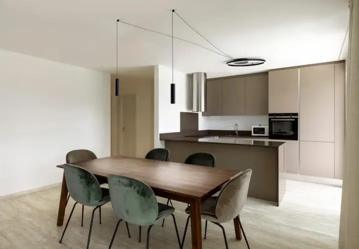 Un lampadario decentrato crea un'atmosfera moderna sopra un tavolo da pranzo in legno con sedie in velluto verde. Soluzioni di illuminazione eleganti per ogni spazio.