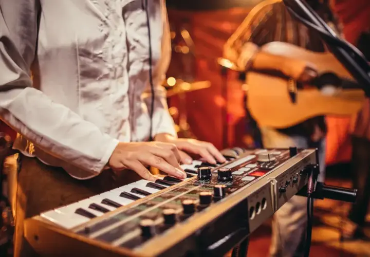 Muzyk gra na keyboardzie, tworząc tło dla piosenek do śpiewania. W tle widać gitarzystę.