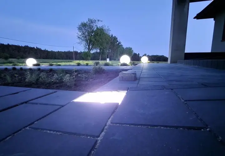 Nocny ogród rozświetlony kulistymi lampami i ledami w kostce brukowej. Ścieżka z ciemnej kostki brukowej prowadzi do domu.