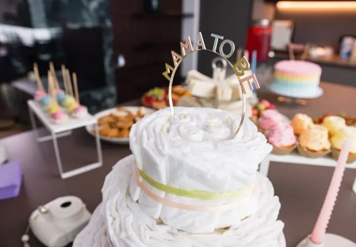 Una torta di pannolini decorata con un topper "Mama to Be" per un baby shower. Sul tavolo anche cupcakes colorati e dolcetti.