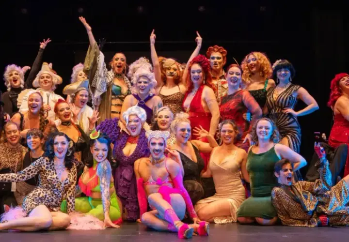 En stor grupp artister i glittrande kostymer poserar glatt på scenen under Stockholm Burlesque Festival.