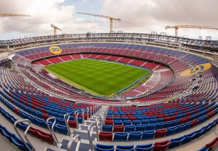 Widok na pusty stadion z czerwono-niebieskimi siedzeniami, w tle widać żurawie budowlane. To jeden z największych stadionów w Polsce, gotowy na przyszłe emocje.