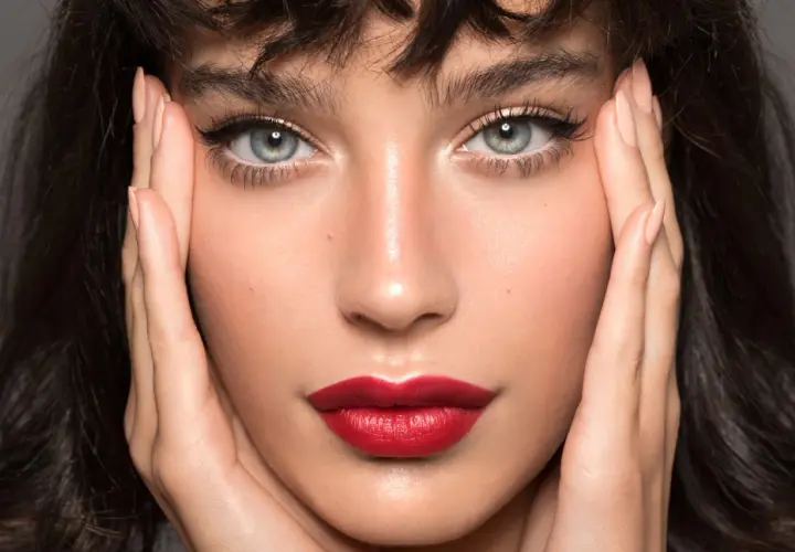 Close-up of a woman's face with perfect winged eyeliner and red lipstick, hands framing her face.