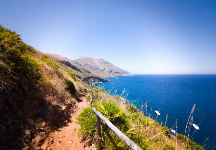 A scenic hiking trail winds through the rugged landscape of Lo Zingaro Nature Reserve, overlooking the brilliant blue Mediterranean Sea dotted with boats.