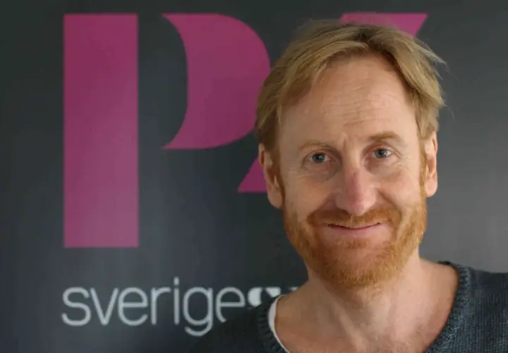 Gustaf Hammarsten, känd från filmer och tv-program, ler mot kameran framför en bakgrund med logotypen för Sveriges Television.