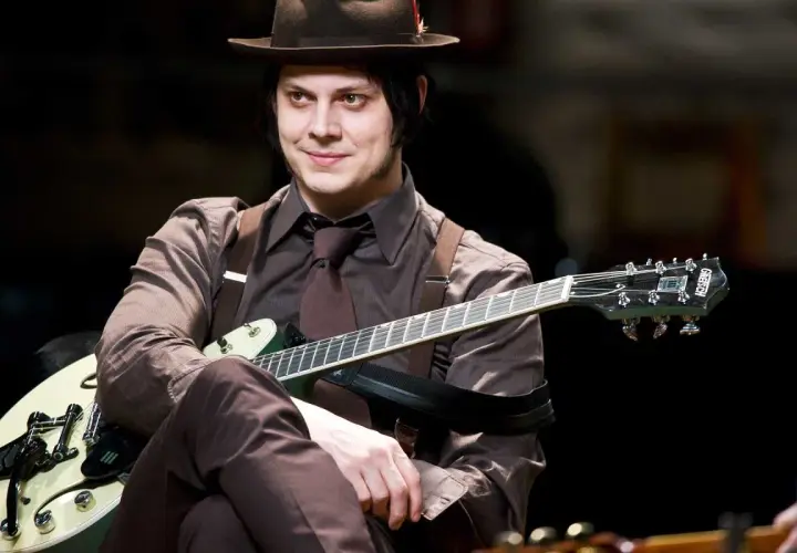 Jack White w brązowym kapeluszu i garniturze gra na gitarze.
