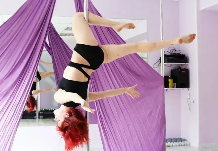 Kobieta z rudymi włosami wykonuje figurę pole dance invert, zwisając z rurki.