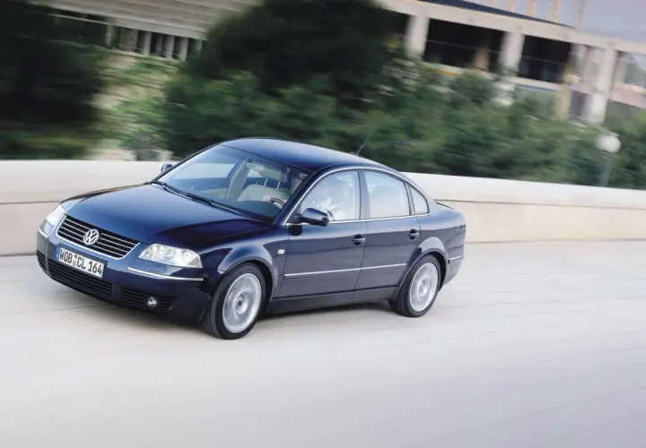 Ciemnoniebieski Volkswagen Passat B5 jedzie po moście, za nim rozmyte drzewa i budynek.