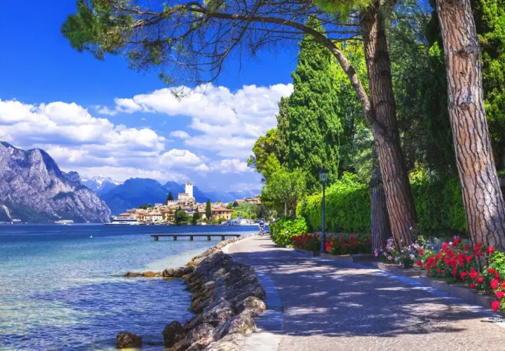 Promenade ensoleillée au bord du lac de Garde, avec des montagnes majestueuses en arrière-plan et un village pittoresque.