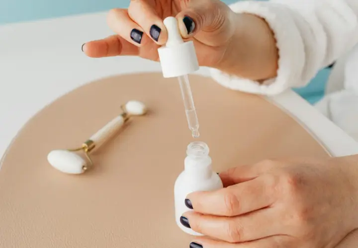 Dłoń z ciemnym lakierem do paznokci trzyma pipetkę z serum, obok jadeitowy roller. Pamiętaj, z czym nie łączyć niacynamidu.