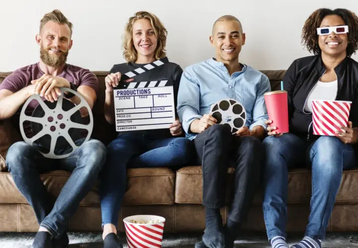 Cztery osoby siedzą na kanapie, trzymając rekwizyty filmowe: klaps, rolki taśmy filmowej, popcorn i napój. Różne rodzaje filmów i ich tworzenie.