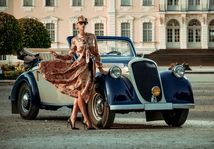 Elegancka kobieta w kwiecistej sukience i okularach przeciwsłonecznych opiera się o zabytkowy kabriolet. Styl lat 20. moda damska.