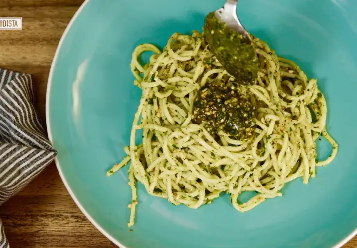 Un plato de pasta al pesto, con una cuchara añadiendo más salsa verde.