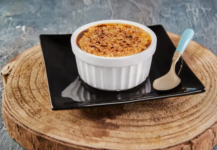 Crème brûlée avec une croûte caramélisée dorée, servie dans un ramequin blanc sur une assiette noire, avec une cuillère à la touche champignon.
