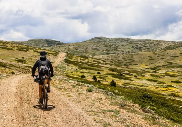Ciclista pedaleando por un camino de tierra en **montañas vacías** bajo un cielo nublado.