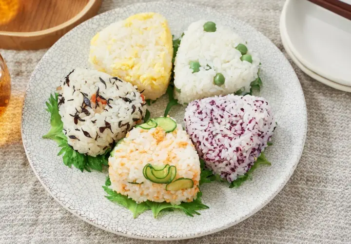 A colorful assortment of musubi vs onigiri, each a unique rice ball shape with various toppings like peas, seaweed, and cucumber.