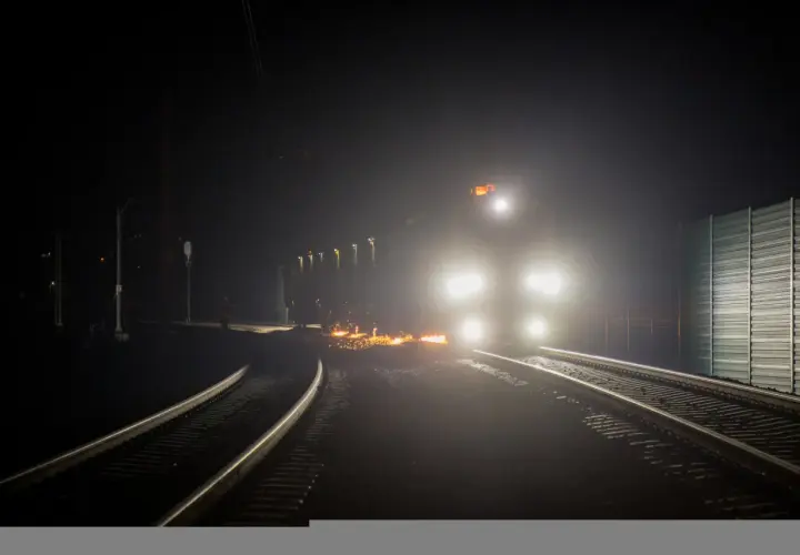 Nocny tor kolejowy rozświetlony reflektorami pociągu i iskrami z prac na torach.