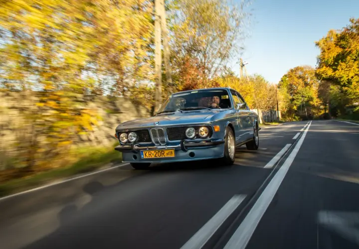 Błękitne BMW E9 z kierowcą jedzie jesienną drogą, otoczone kolorowymi drzewami.