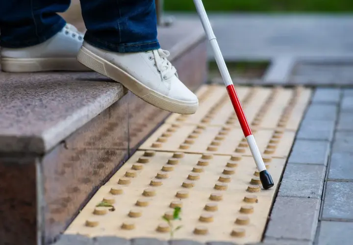 Osoba w białych butach i jeansach używa białej laski z czerwonym paskiem na żółtej nawierzchni z wypustkami.