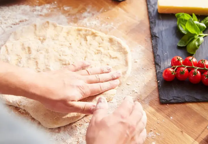 Hände formen den **pizzateig auftauen** auf einer bemehlten Holzoberfläche. Daneben liegen frische Tomaten, Mozzarella und Basilikum.
