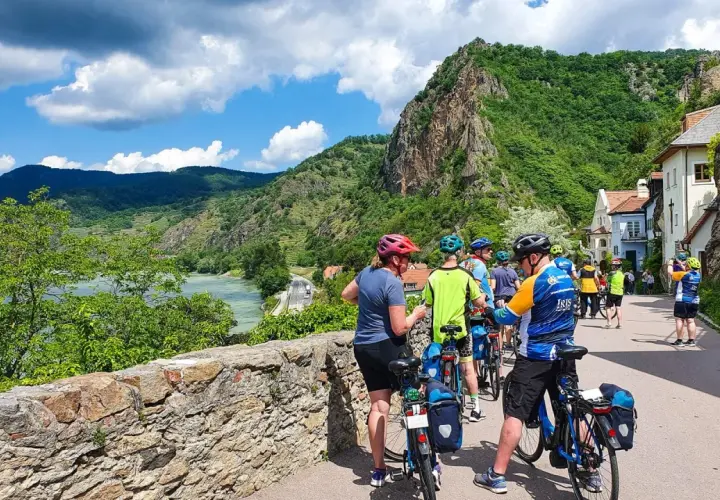 A group of cyclists on a Danube bike tour pauses by a stone wall, with a river and lush green hills in the background.