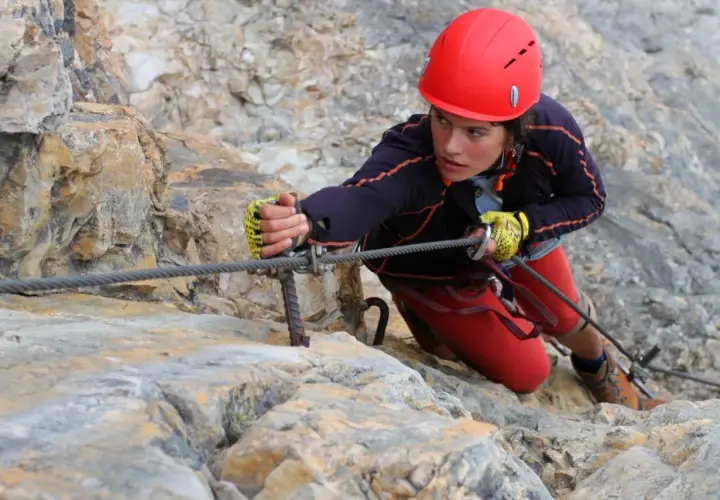 Młoda kobieta w kasku wspina się po skale, używając stalowej liny. To przygoda na via ferrata Czechy.