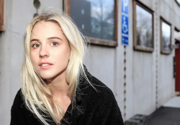 En ung kvinna med blont hår, Paolo Nobis Sandén, tittar in i kameran framför en grå byggnad med fönster.